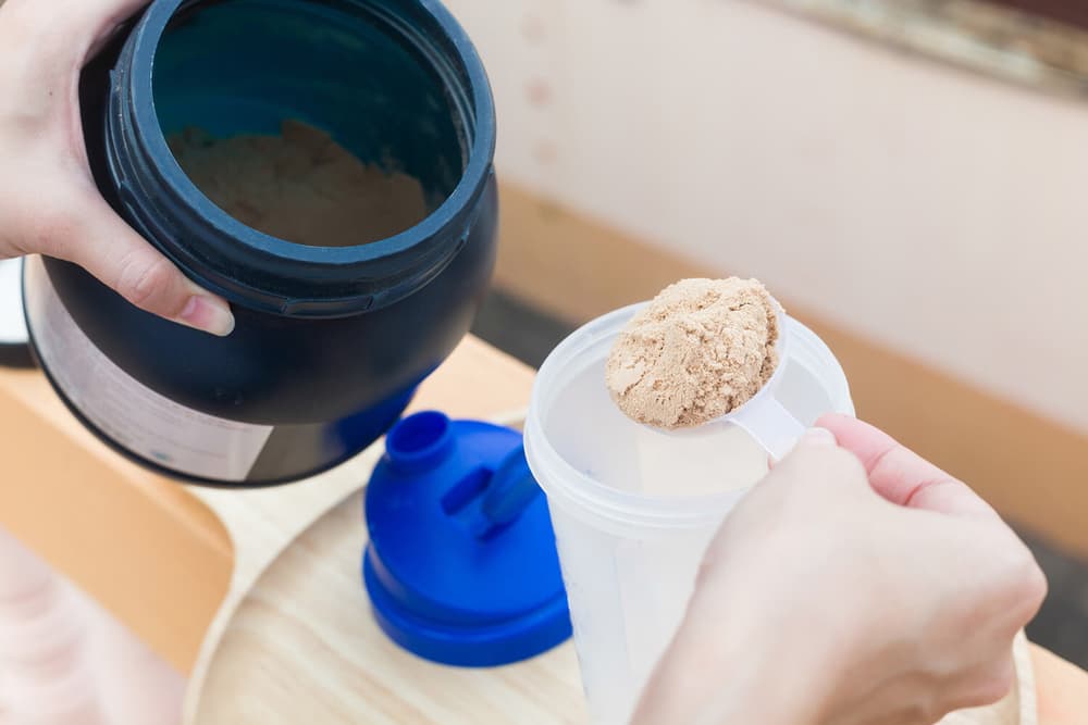 A person scoops protein powder from a black container into a clear shaker bottle with a blue lid.