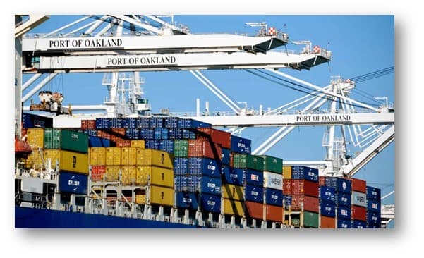 Cargo containers are stacked on a ship at the Port of Oakland, with large white cranes labeled Port of Oakland visible in the background under a clear blue sky.
