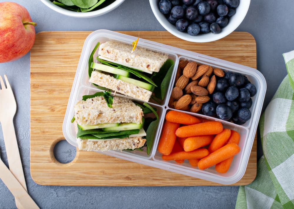 A lunchbox with cucumber sandwiches, baby carrots, almonds, and blueberries on a wooden board. A bowl of blueberries and an apple are nearby.
