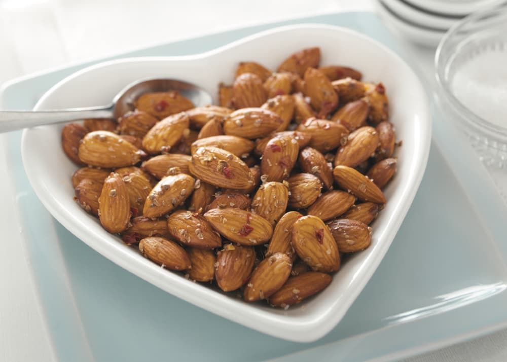 Heart-shaped bowl filled with roasted almonds, seasoned with herbs and red chili flakes, with a small spoon resting on the edge.
