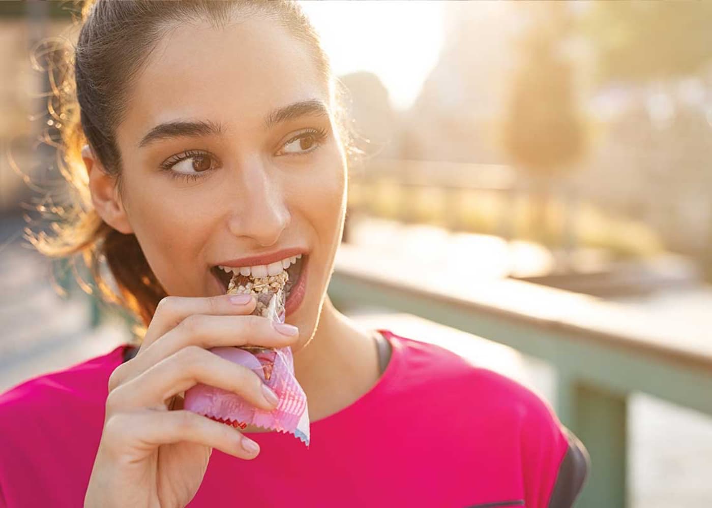 A person wearing a bright pink athletic top eats a granola bar outdoors in sunlight.