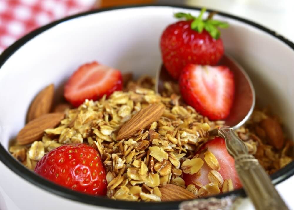 A bowl of granola with whole almonds and sliced strawberries, with a spoon resting inside.