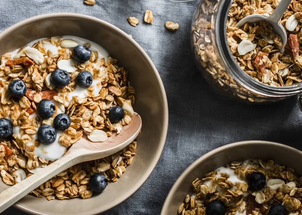 Bowls of granola topped with blueberries and yogurt, with a wooden spoon and a jar of granola on a gray surface.