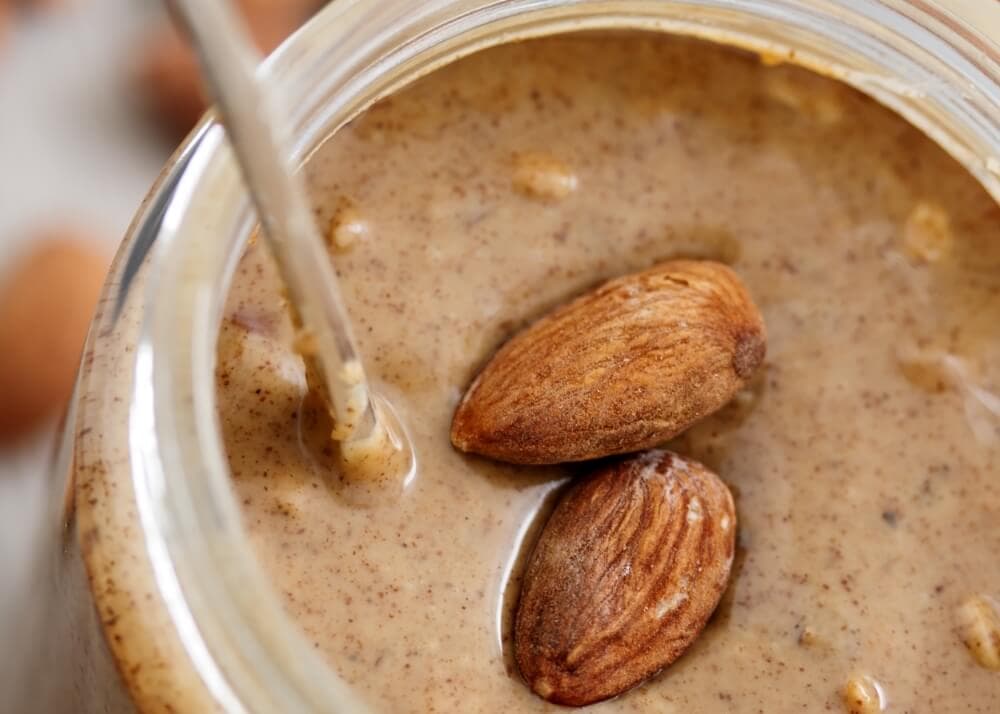 Close-up of a jar of almond butter with two whole almonds on top and a metal spoon partially visible on the left side.
