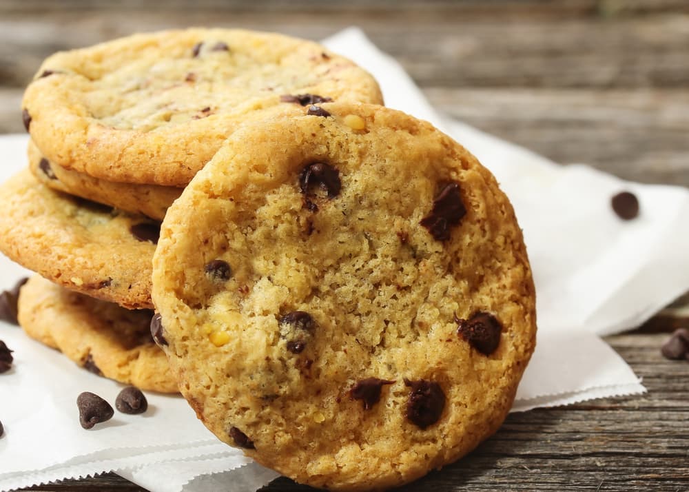 A stack of chocolate chip cookies rests on white parchment paper, with scattered chocolate chips on a wooden surface in the background.