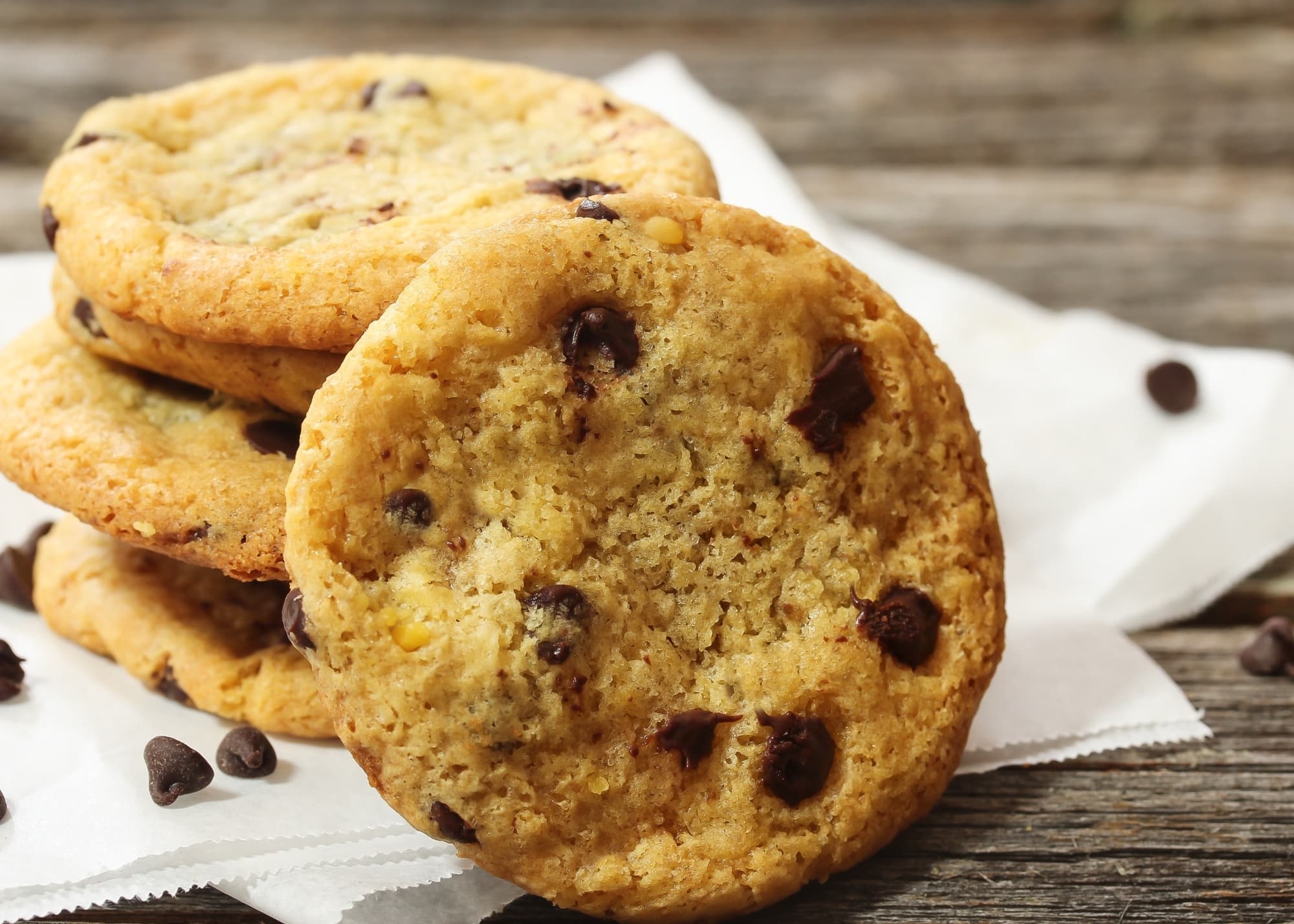 A stack of chocolate chip cookies rests on white parchment paper, with scattered chocolate chips on a wooden surface in the background.
