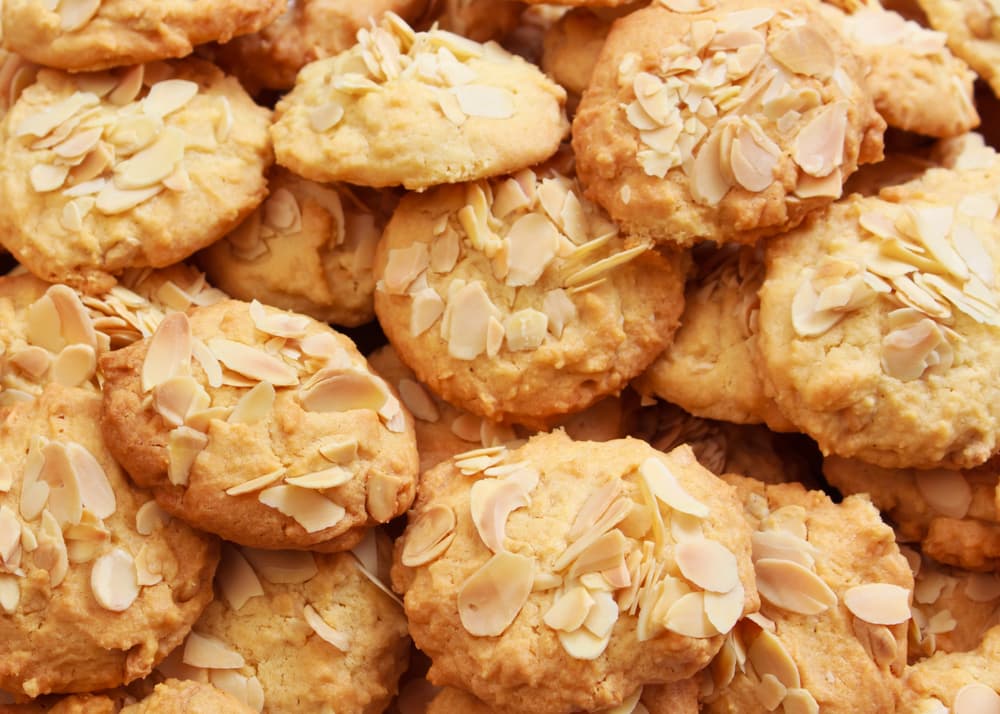A pile of round almond cookies, each topped with sliced almonds, closely arranged and overlapping on a flat surface.
