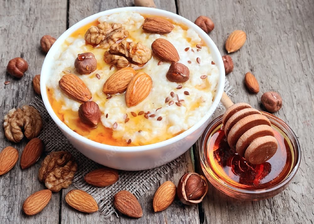 A bowl of oatmeal topped with walnuts, almonds, hazelnuts, flaxseeds, and honey sits on a wooden surface next to a small dish of honey and a honey dipper.