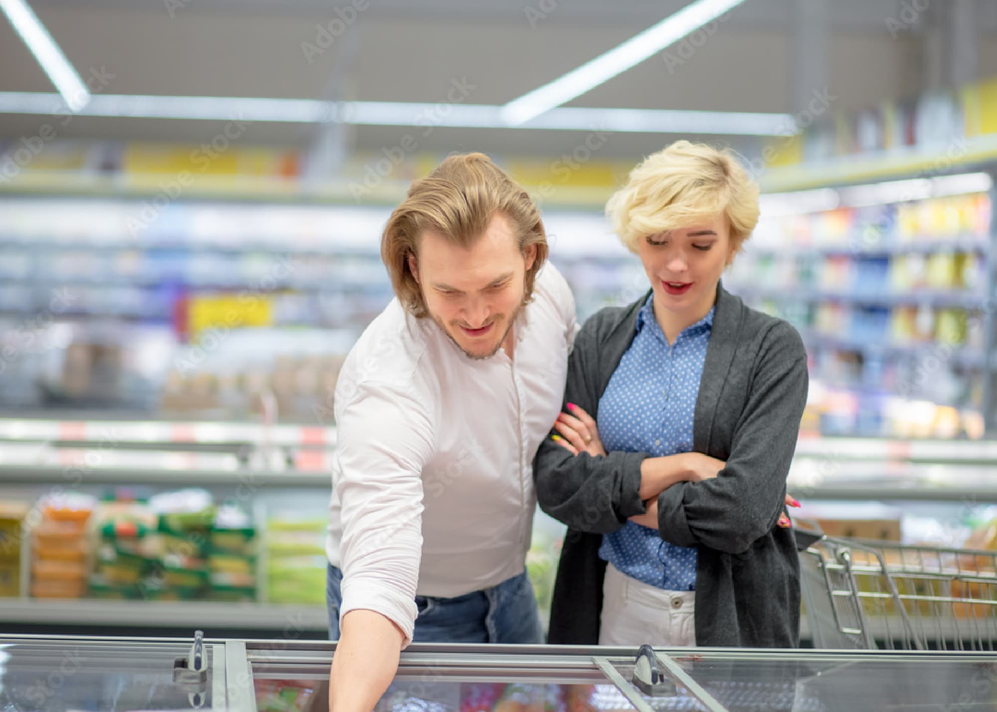 A man and woman stand by a freezer in a grocery store, looking at frozen food options together. The man is reaching inside while the woman watches with her arms crossed.