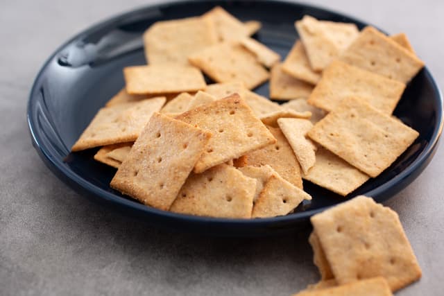 A dark plate with several square, golden brown crackers scattered on it, with a few crackers on the table beside the plate.