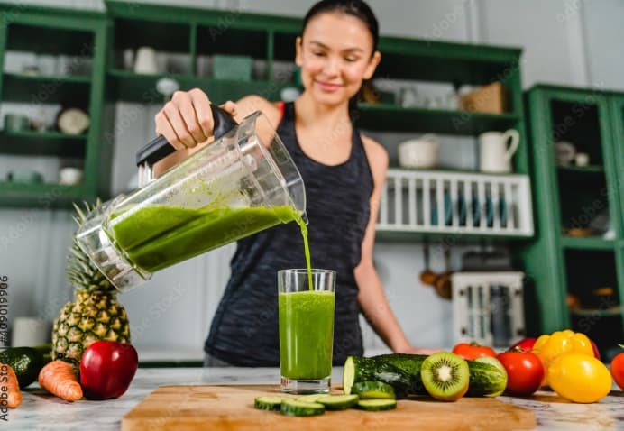 A woman pours green smoothie from a blender into a glass on a kitchen counter with assorted fresh vegetables and fruit.