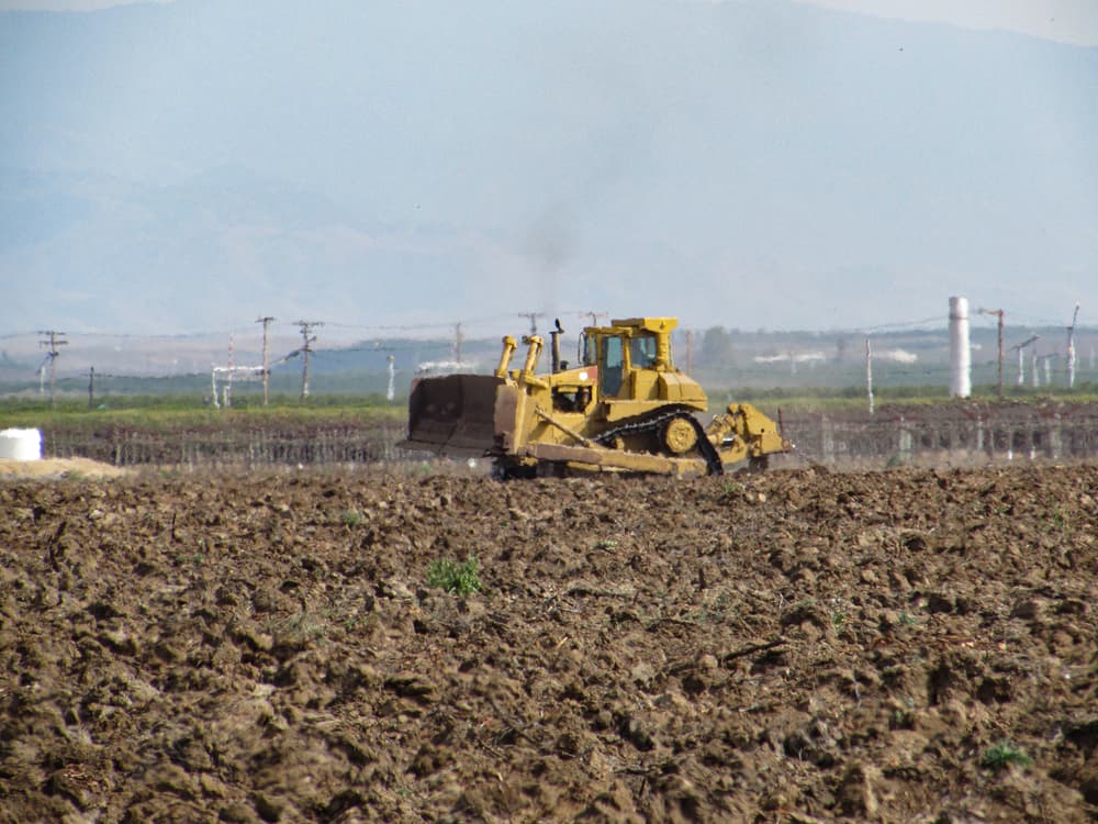 A yellow bulldozer is pushing soil across a plowed field with power lines and distant mountains in the background.