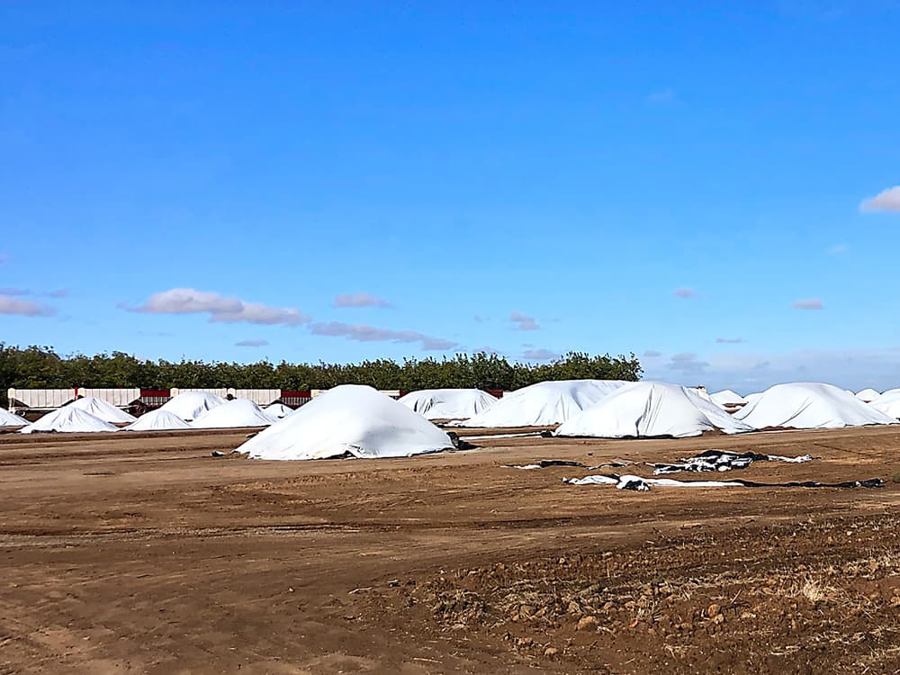 Several large piles of material covered with white tarps sit on bare dirt under a blue sky, with trees and a long row of train cars in the background.
