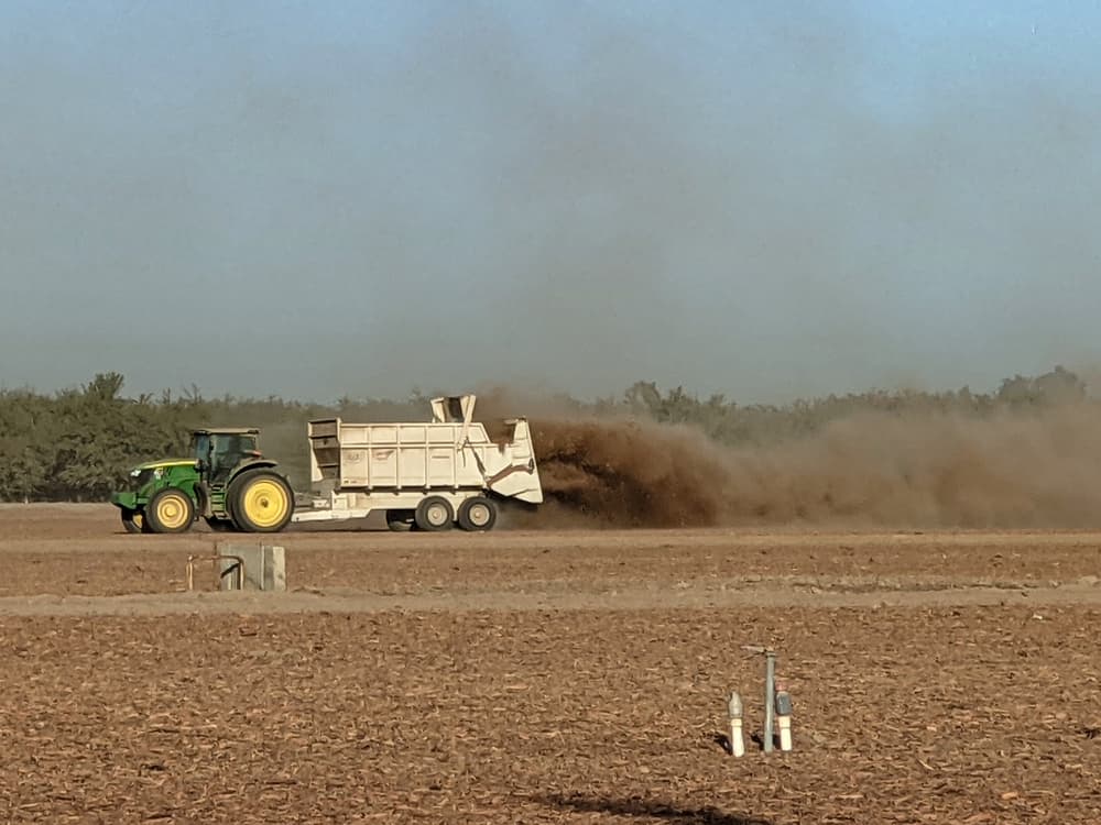 A green tractor pulls a white trailer spreading a cloud of material over a dry, empty field under a clear blue sky.