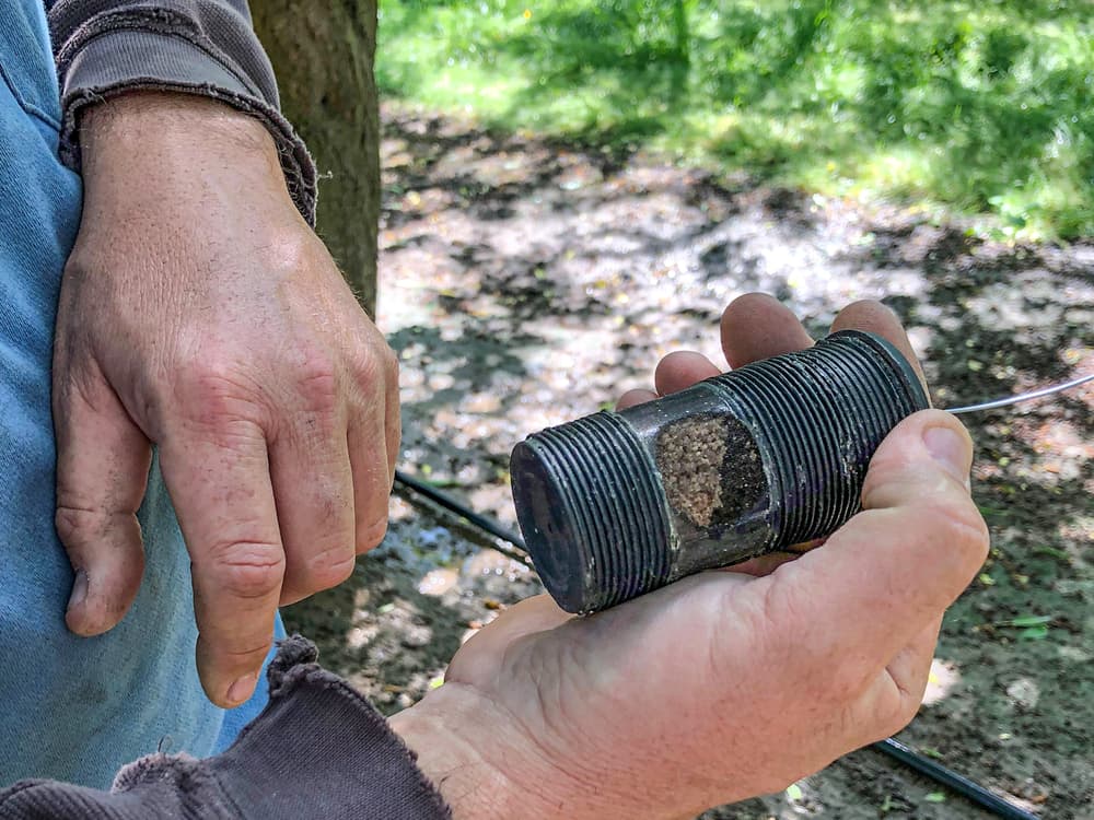 Two hands hold a metal pipe section with threaded ends and corrosion or mineral buildup inside, outdoors on a sunny day.