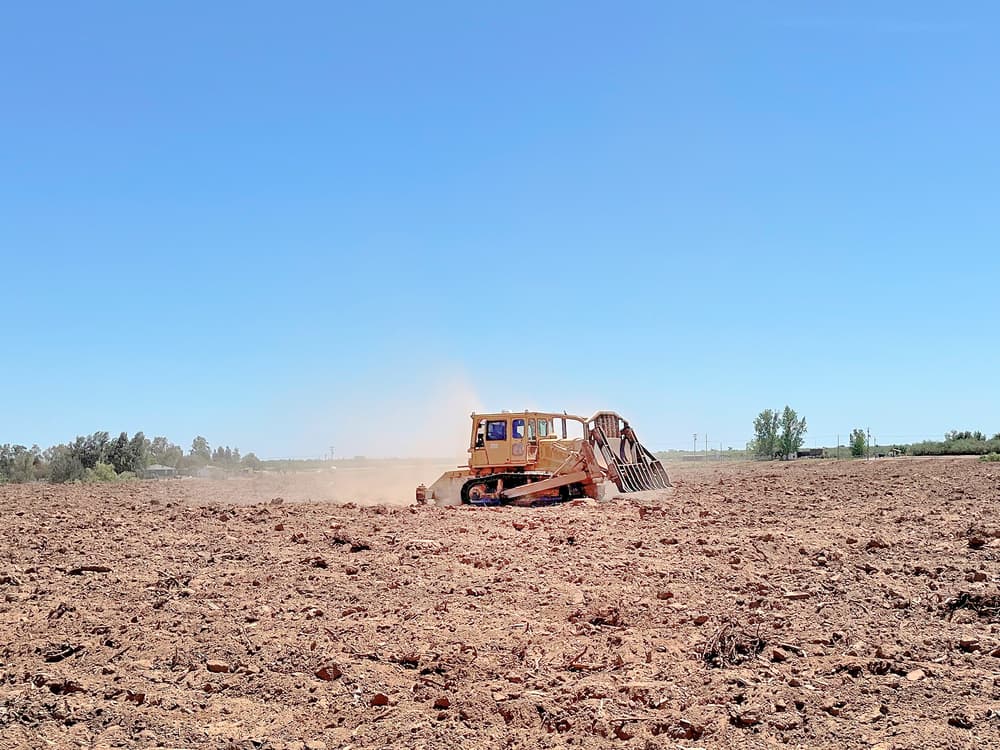 A yellow tractor with a large attachment works on a dry, plowed field under a clear blue sky, raising dust as it moves. Trees and bushes are visible in the background.