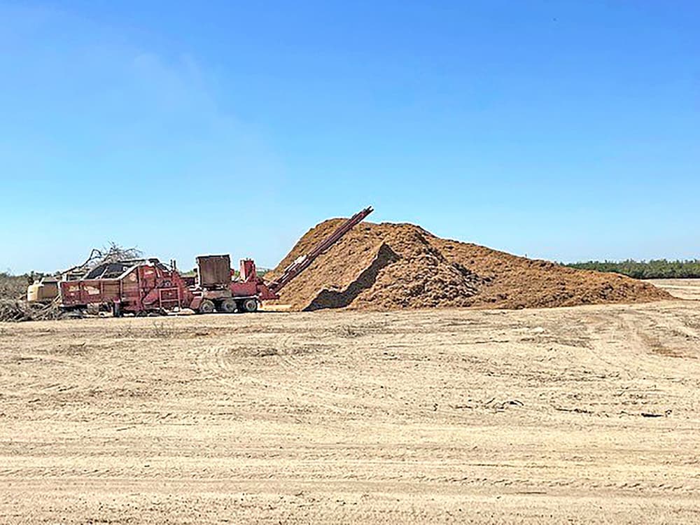 A large pile of mulch sits on a dirt lot with industrial equipment nearby under a clear blue sky.