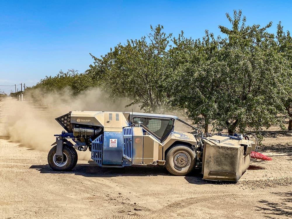 A large, dust-covered agricultural machine operates on a dirt path between rows of trees under a clear blue sky.
