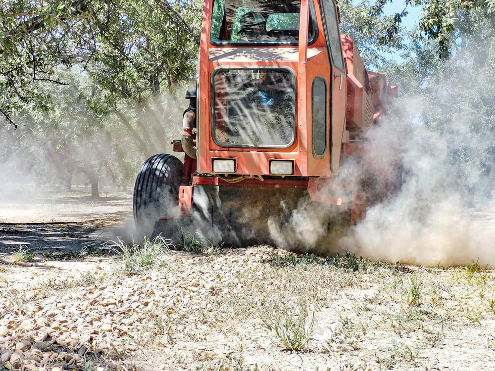 A red agricultural machine moves through a field, creating dust as it processes or collects crops from the ground under trees.