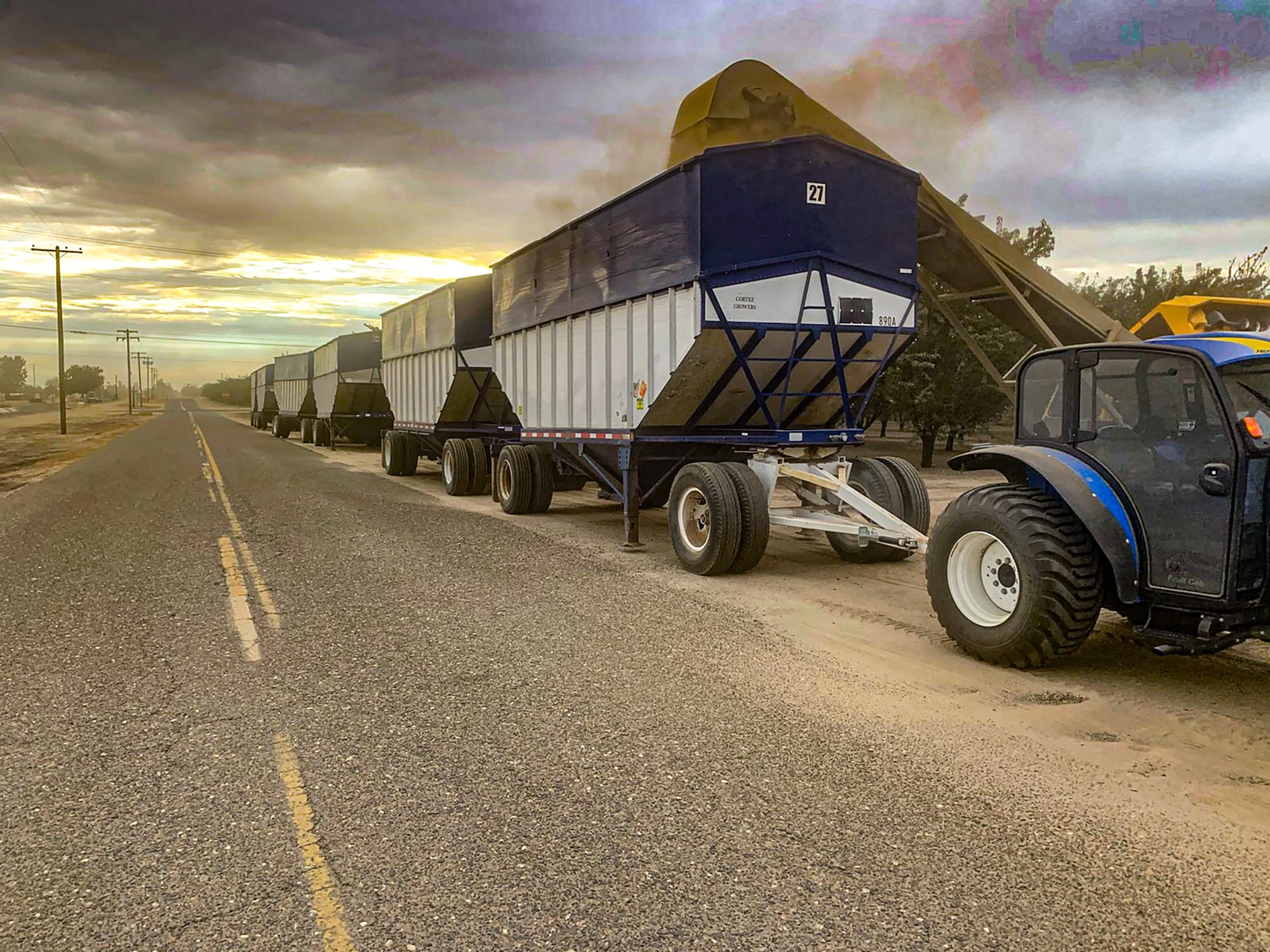 A row of large trailers is parked on the roadside, with a tractor unloading or loading material at sunrise under a cloudy sky.