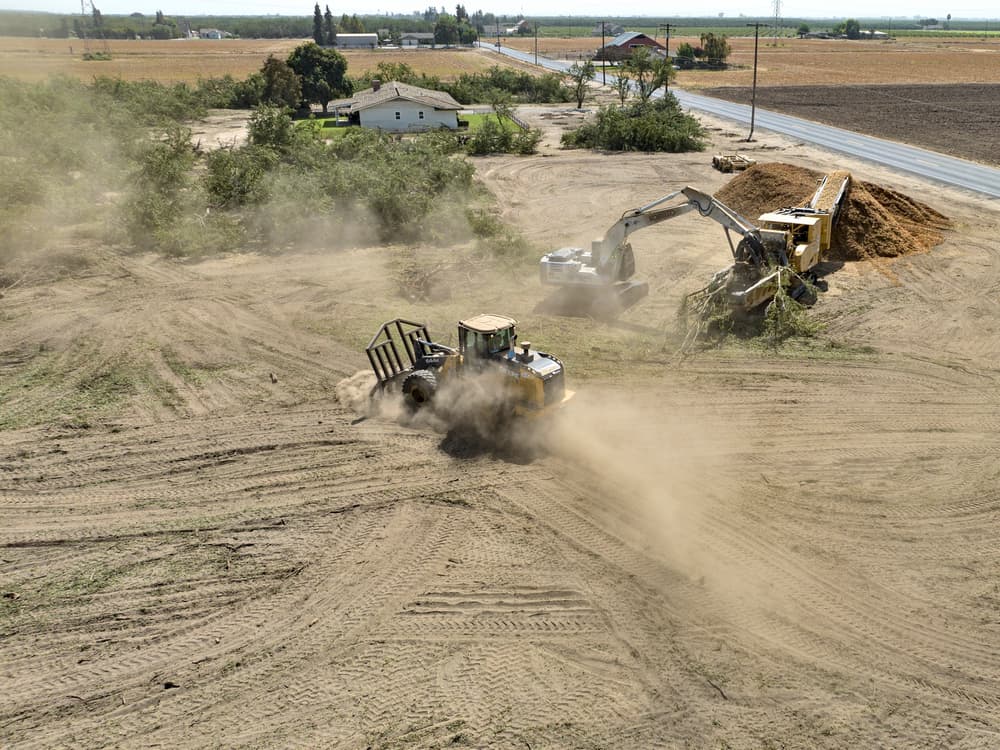 A bulldozer and an excavator move dirt and create dust at a construction site next to a road in a rural area.