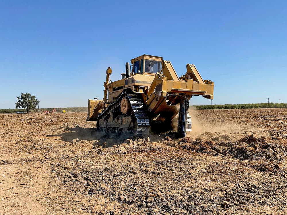 Large yellow bulldozer with metal tracks moves dirt on a cleared, dry field under a clear blue sky.