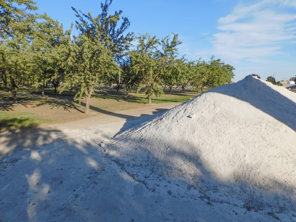 A large pile of white sand or gravel sits near trees in an outdoor area under a clear blue sky.