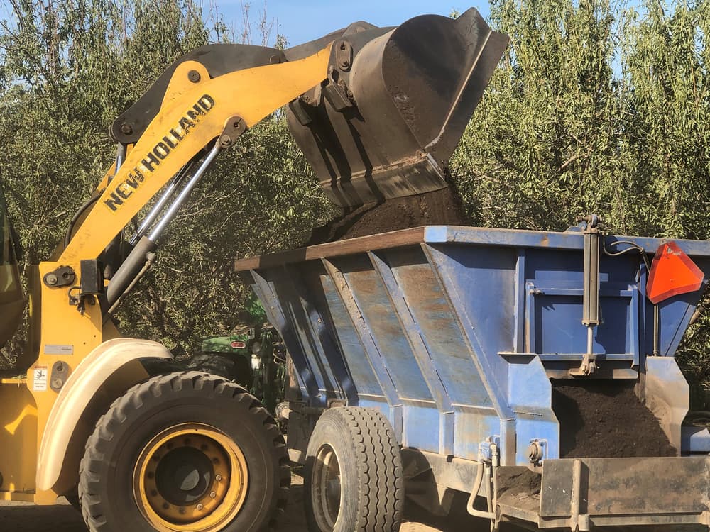 A yellow New Holland loader pours soil or compost into a large blue trailer outdoors, with trees in the background.