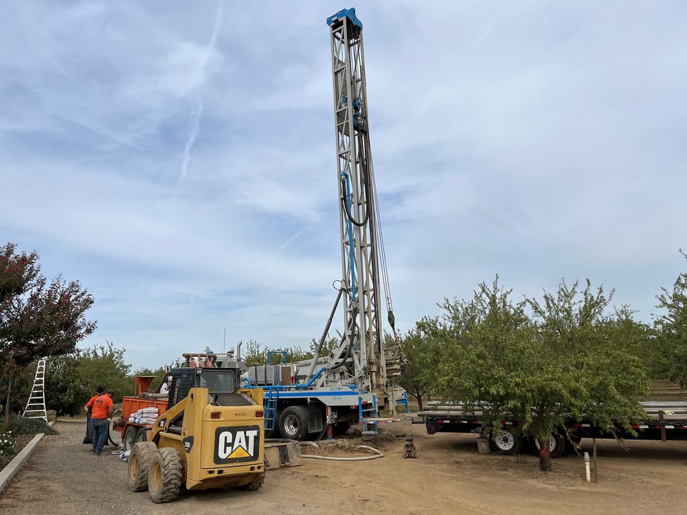 A drilling rig operates in a rural area with workers nearby, a yellow CAT loader in the foreground, and trees surrounding the site.