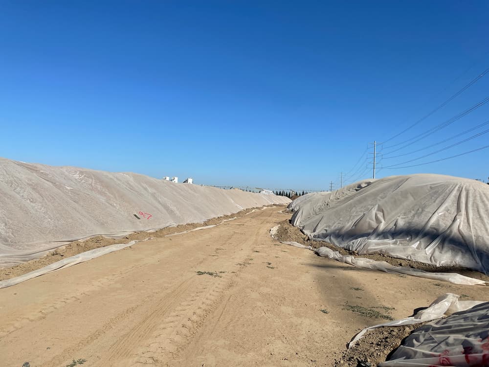 Dirt road flanked by large piles of sand or soil covered with plastic sheets, under a clear blue sky with power lines running alongside.