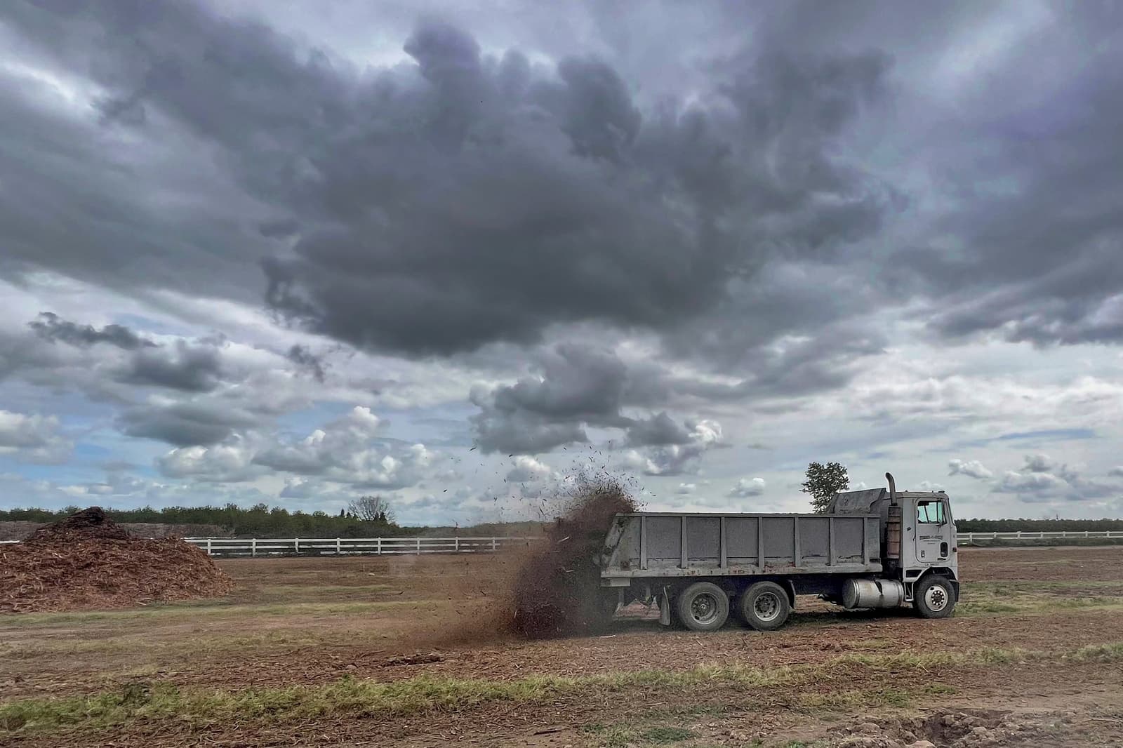 A dump truck unloads a pile of dirt onto a field under a cloudy sky, with a white fence and trees in the background.