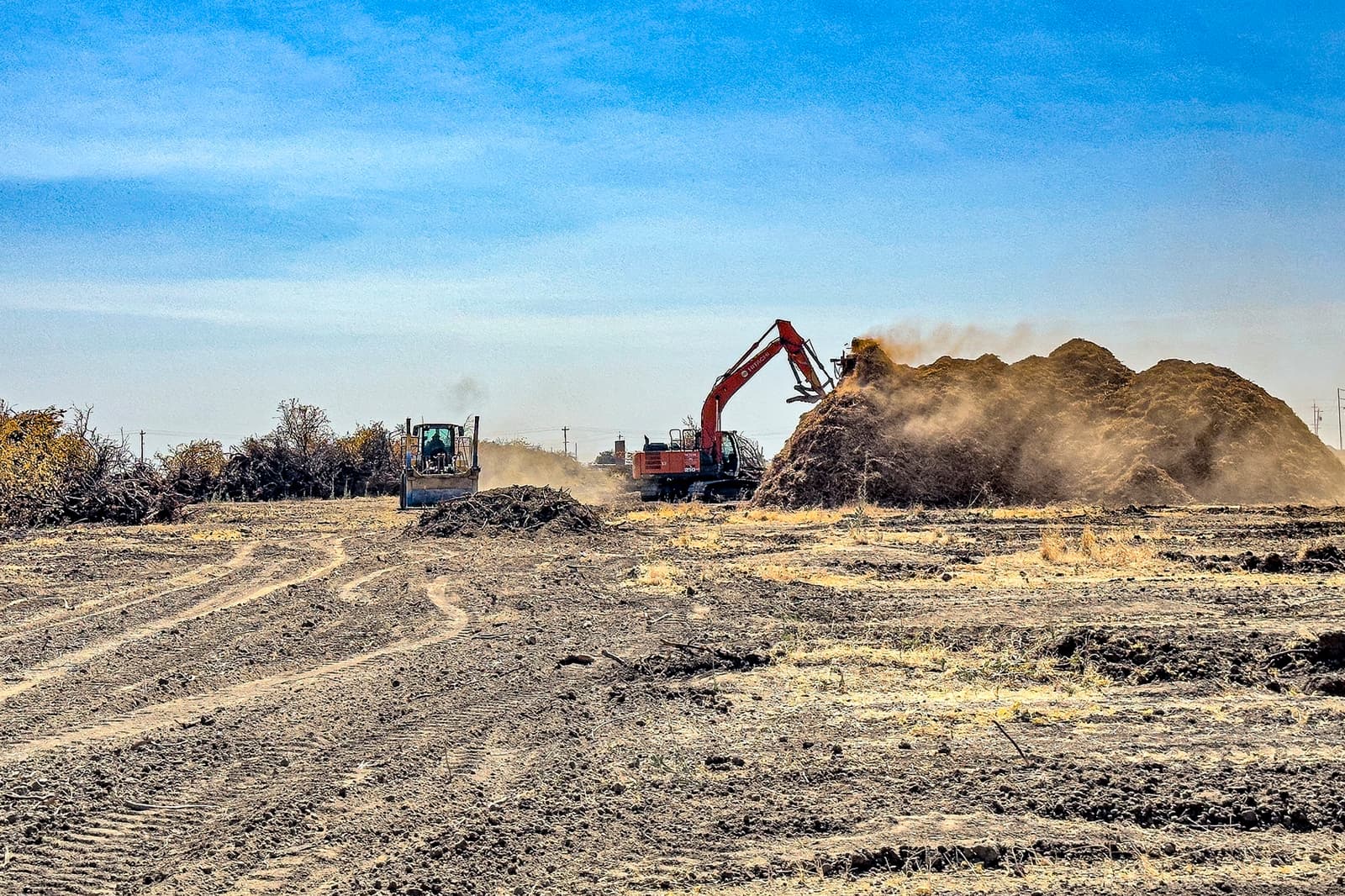 Excavator moves a pile of dirt while a bulldozer works nearby on a cleared, open construction site under a clear blue sky.