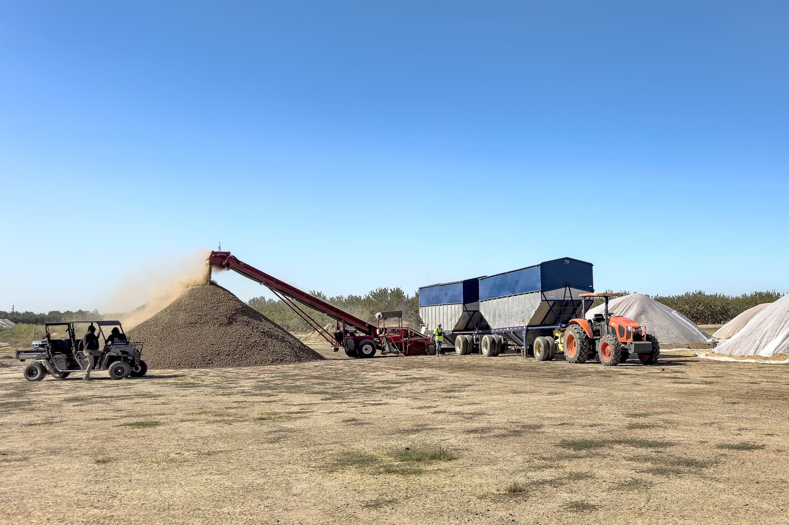 A conveyor unloads material onto a large pile beside a trailer and a tractor; a utility vehicle is parked nearby under a clear sky.