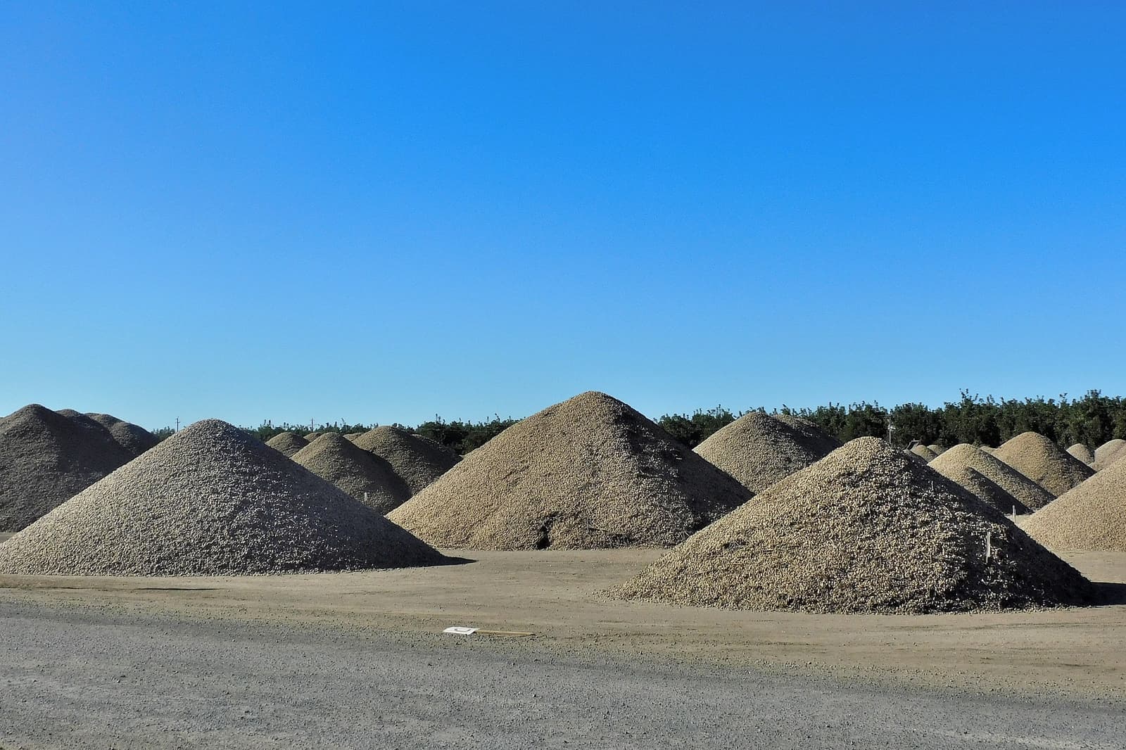 Large piles of gravel are arranged in rows on a dirt lot under a clear blue sky, with trees visible in the background.