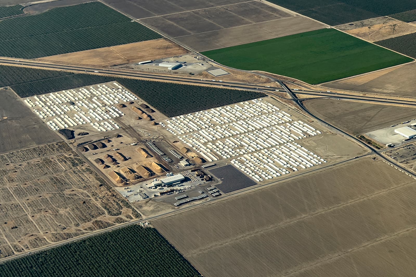Aerial view of a large industrial facility with rows of white structures, surrounded by agricultural fields, roads, and some buildings.