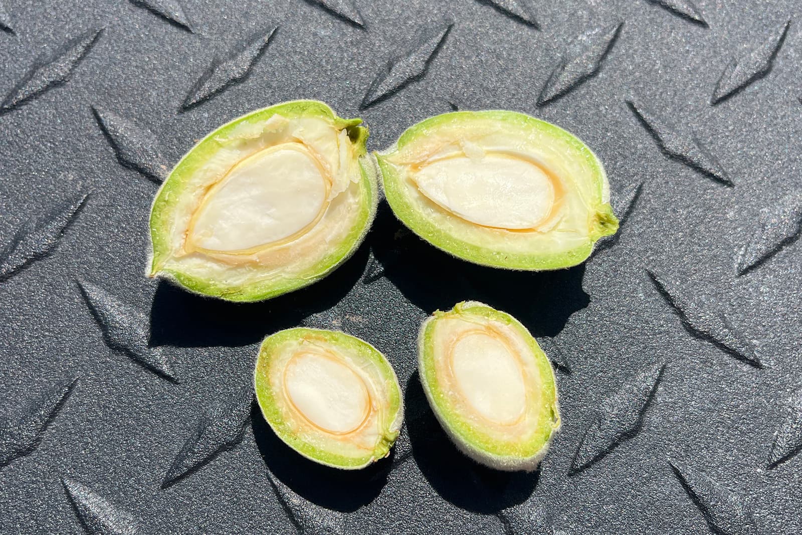Four green almonds, two whole and two halved, are placed on a textured black surface with a diamond plate pattern, showing their white interior and seed.