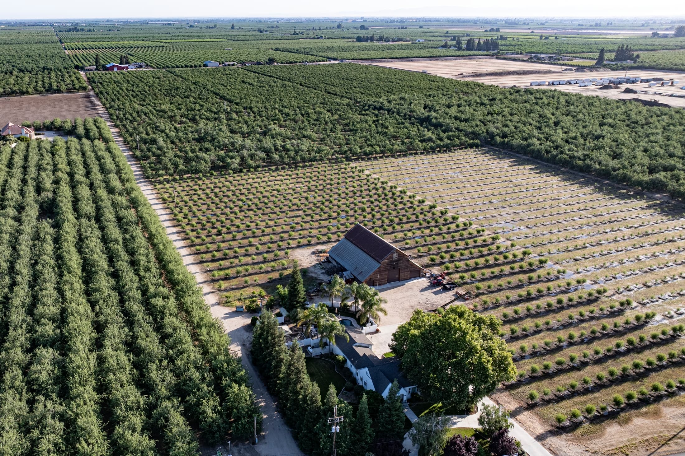 Aerial view of a large farm with rows of crops and orchards, a barn, and farmhouse surrounded by trees in a rural landscape.