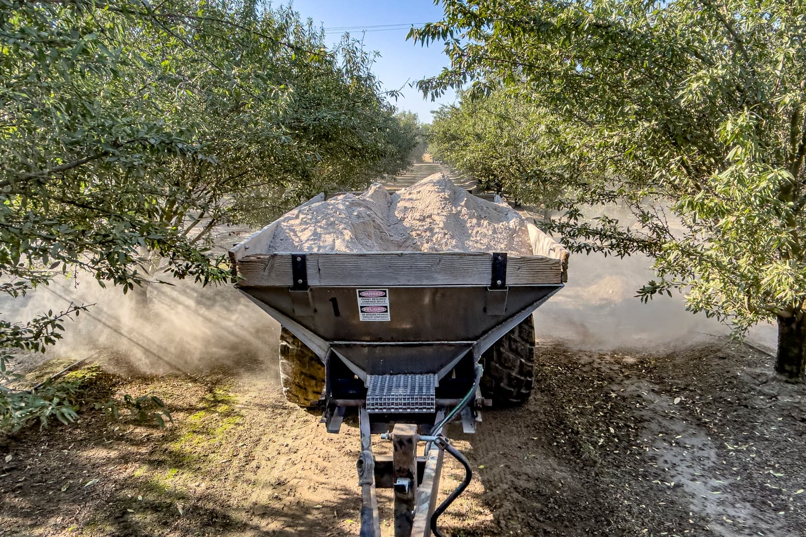 A tractor pulls a trailer filled with soil or sand down a dirt path between rows of trees, raising dust clouds along the way.