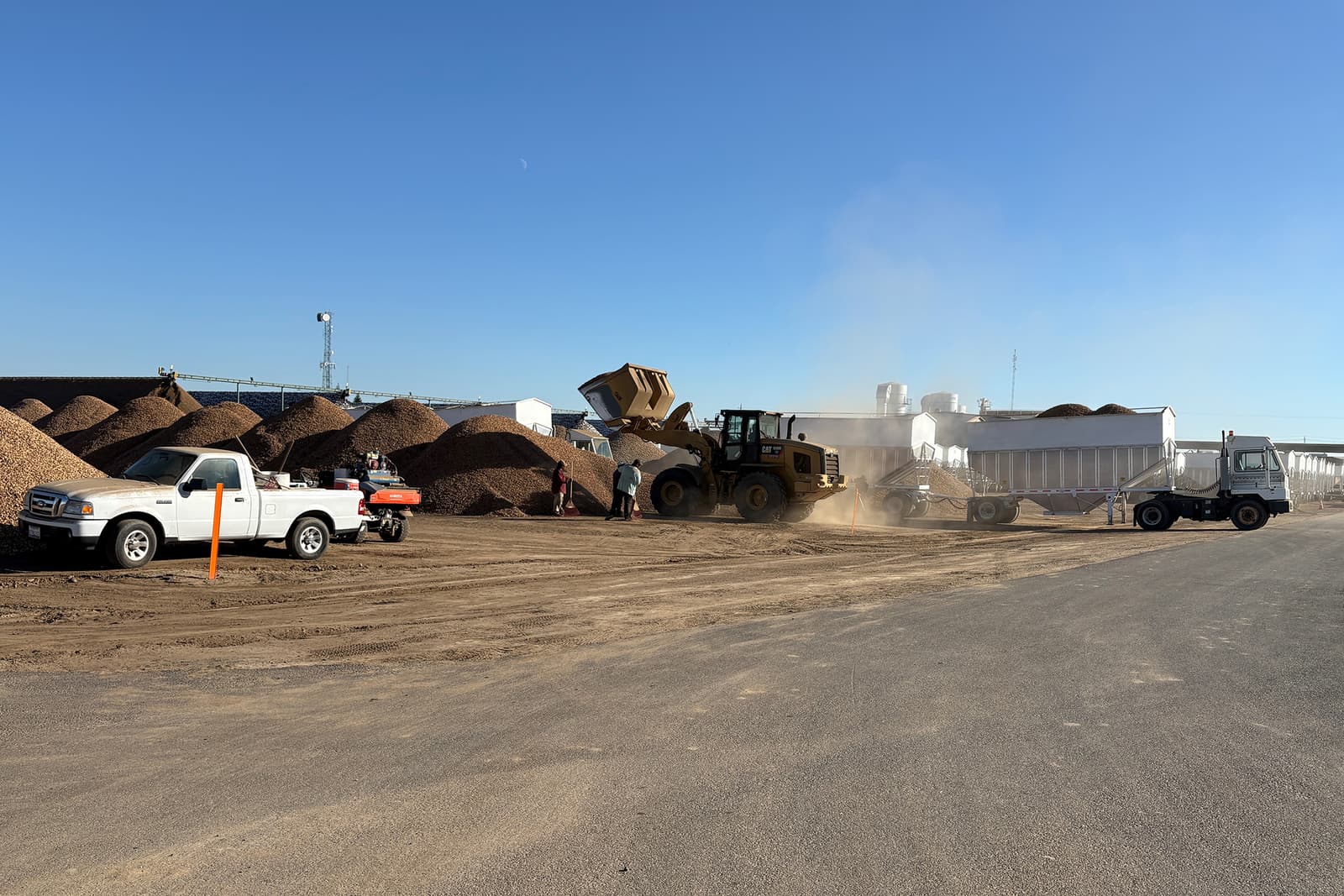A loader dumps material into a truck trailer near piles of dirt or gravel, with two parked vehicles nearby on a clear sunny day.