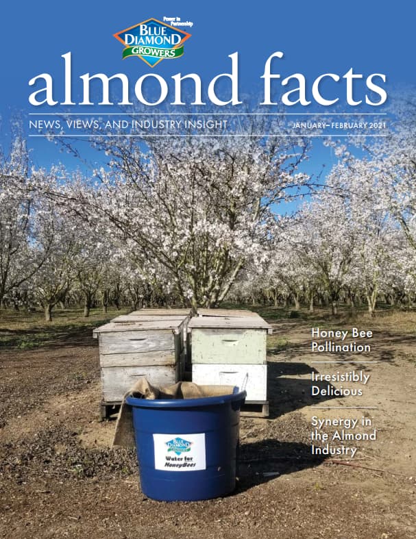 Beehives and a blue bin labeled Whole Bee Health Manufacturer sit among blooming almond trees under a clear sky; magazine cover text includes headlines about honey bee pollination and the almond industry.