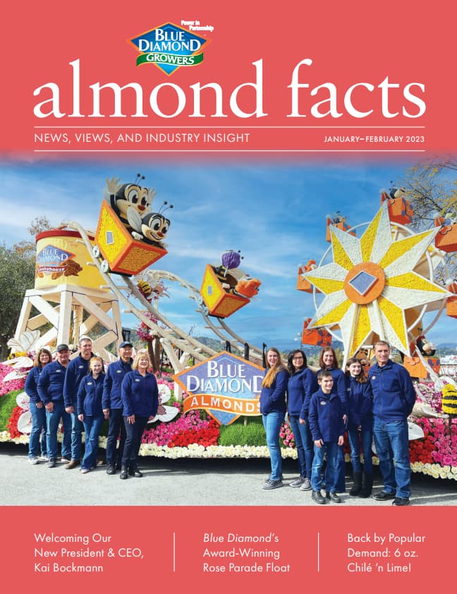A group of people in blue jackets pose in front of a colorful Blue Diamond Growers almond-themed parade float with cartoon characters and large decorative elements.