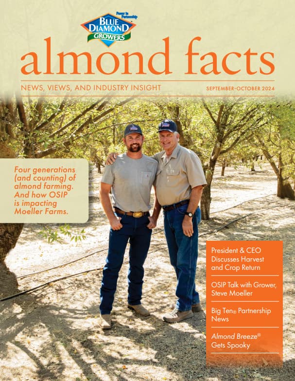 Two men stand together in an almond orchard under tree shade, both wearing jeans, light shirts, and hats, posing for the cover of a farming industry magazine.