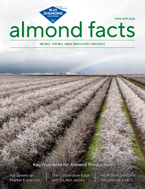 A blooming almond orchard stretches into the distance under a cloudy sky with a faint rainbow, featured on the cover of almond facts magazine, March-April 2026 issue.