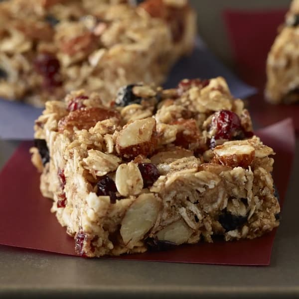 A close-up of a homemade granola bar with visible oats, nuts, and dried fruit, resting on a piece of maroon paper.