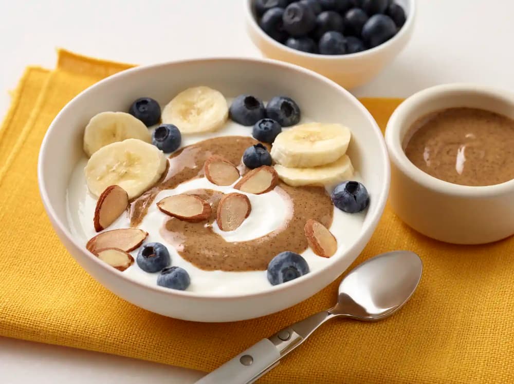Bowl of yogurt topped with banana slices, blueberries, almond butter, and almond slices, with a spoon, napkin, extra blueberries, and almond butter nearby.