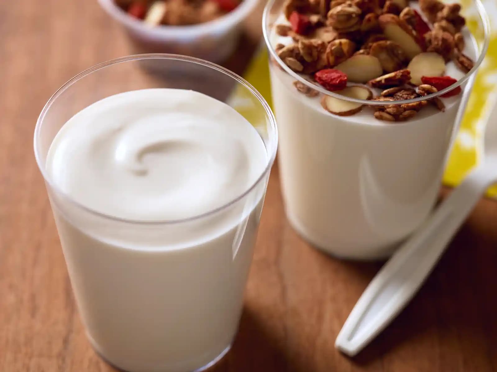 Two clear cups on a wooden table, one filled with plain yogurt, the other filled with yogurt topped with granola and dried fruit. A spoon and another small cup are in the background.