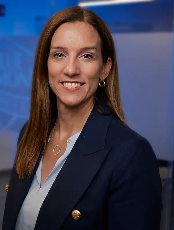A woman with straight brown hair, wearing a navy blazer and light blue blouse, smiles at the camera against a blue-toned office background.